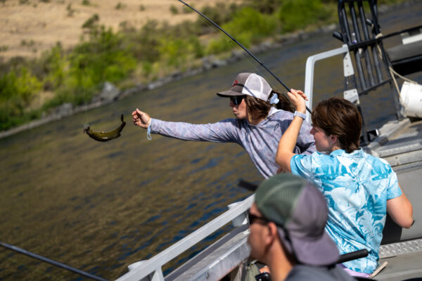 People fish from a boat as a small fish hangs from a fishing line above the river.