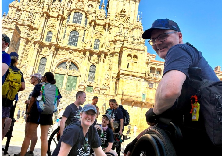 Visitors pose in front of an ornate sunlit cathedral, including a smiling man seated in a wheelchair.