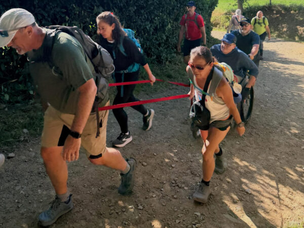 A group of hikers pulls a person in an all-terrain mobility chair along a rocky dirt trail using red straps.