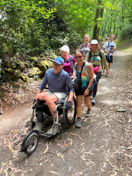 Group of hikers on a forest trail, including a man in an adaptive wheelchair and companions walking alongside him.