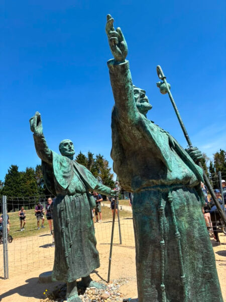 Low-angle view of two large weathered bronze statues with raised arms in a sandy outdoor area, with a volleyball net and people in the background.