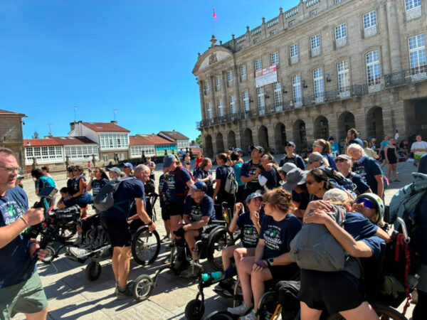 A large group, including wheelchair users, gathers in a sunny plaza before an ornate historic stone building.