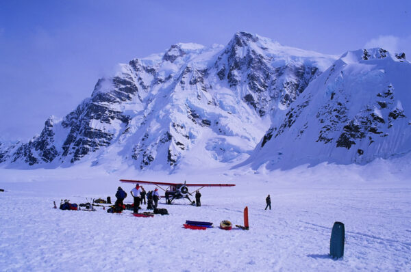 A small red-and-white ski plane and several people on a glacier beneath a towering, snow-covered mountain.