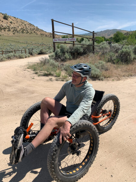 Person seated in an orange adaptive off-road cycle on a dirt path beneath a “No Through Traffic” sign.