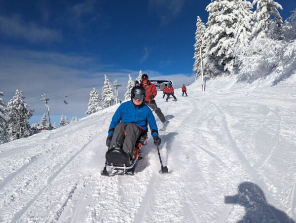 A person uses an adaptive sit-ski while descending a snow-covered slope, with other skiers, a ski lift, and snow-covered trees in the background.