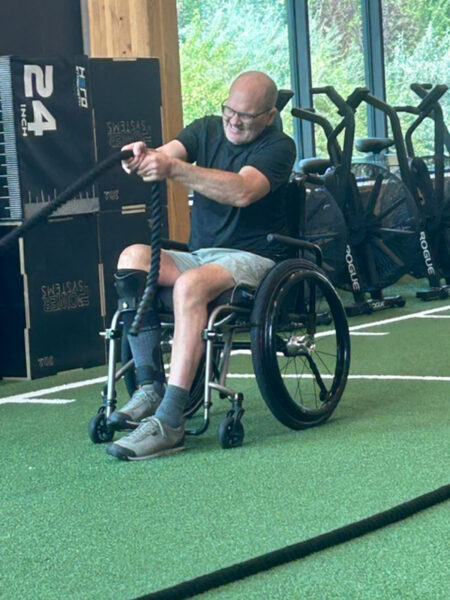 Person in a wheelchair performing a rope-pulling exercise in a gym.