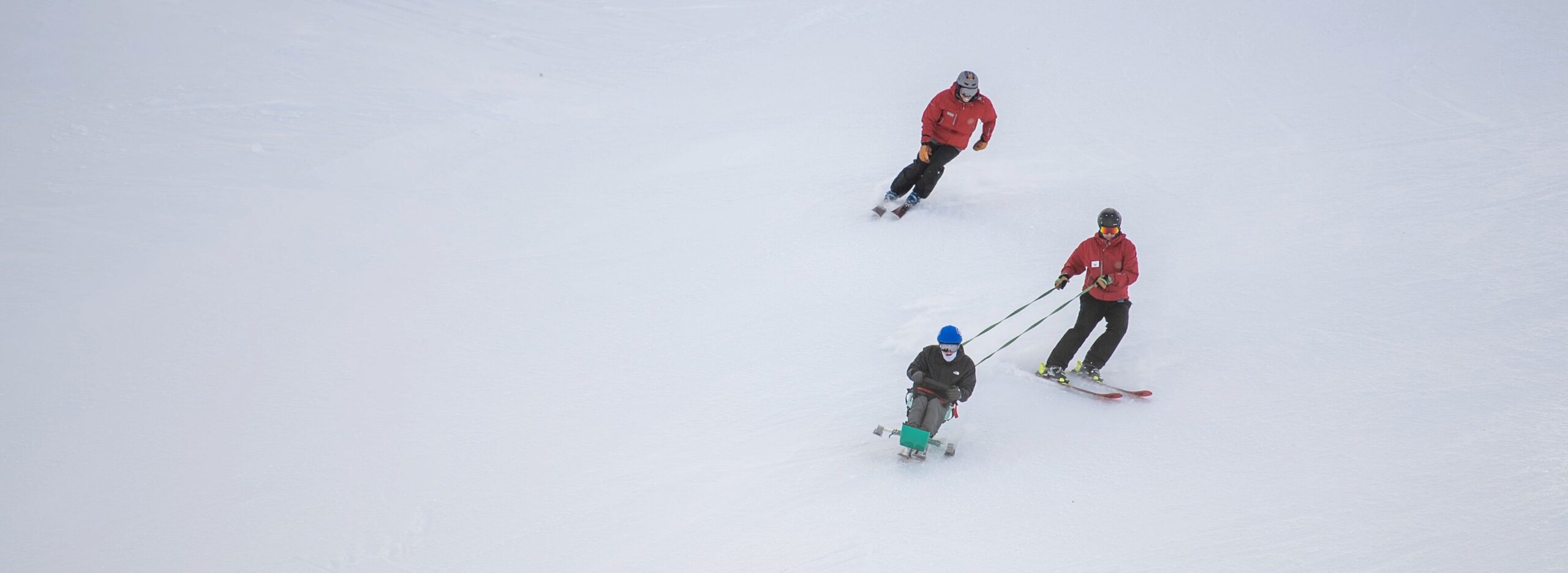 Three people ski downhill on a snowy slope, with one skier pulling a seated person in an adaptive ski device.