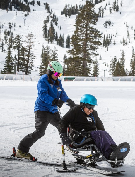Two people participate in adaptive skiing, with an instructor guiding a seated skier in a sit-ski on a snowy slope.