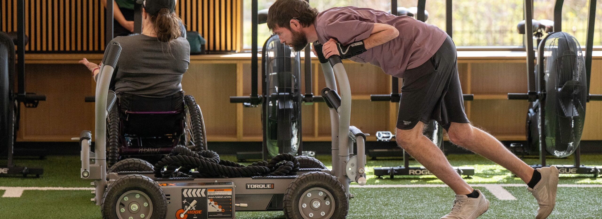Two people exercise with a wheeled resistance sled in an indoor gym; one person uses a wheelchair while another pushes the sled.