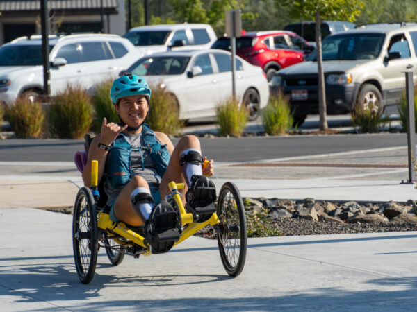 A young adult smiling and riding a yellow handcycle, giving a "shaka" gesture, in an outdoor environment with parked cars in the background.