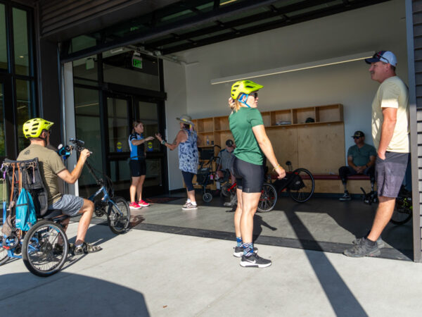 A group of individuals, including people on adaptive bikes, gathers outside a recreational facility, engaging in conversation and social interaction.
