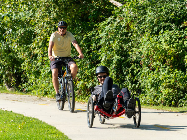 Two cyclists on a path: one on an upright bicycle, the other on a recumbent bike, both surrounded by green foliage.