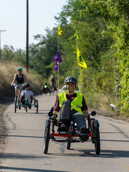 Cyclists riding recumbent tricycles on a path, with one person smiling in a yellow vest at the forefront.