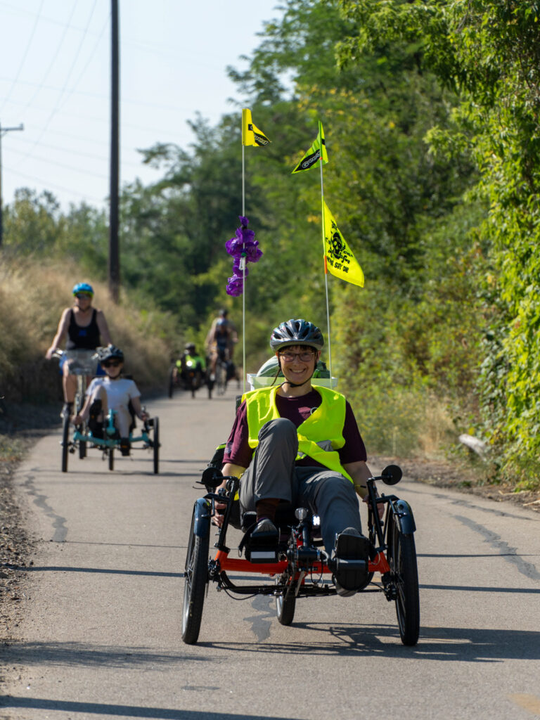 Cyclists riding recumbent tricycles on a path, with one person smiling in a yellow vest at the forefront.