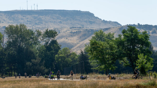 A park scene with people cycling and jogging along a pathway, framed by trees and rolling hills in the background, topped with communication towers.