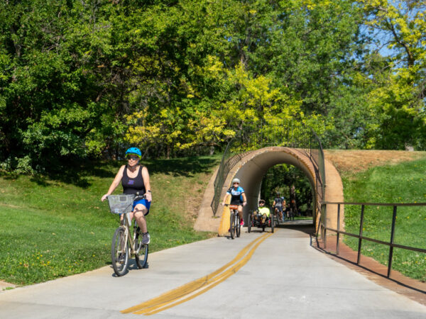 A woman in a blue helmet rides a bike on a paved path with a rounded tunnel ahead. Other cyclists follow along a lush green trail.