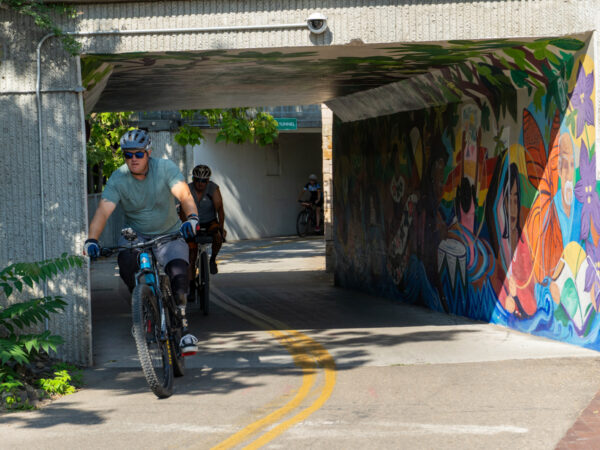 Two cyclists riding through a concrete tunnel adorned with a colorful mural, surrounded by greenery.