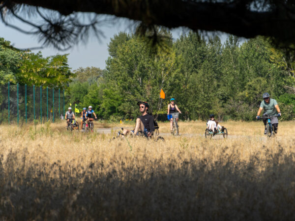 A group of cyclists, including adults and a child, ride along a dirt path surrounded by dry grass and green trees in a sunny outdoor setting.