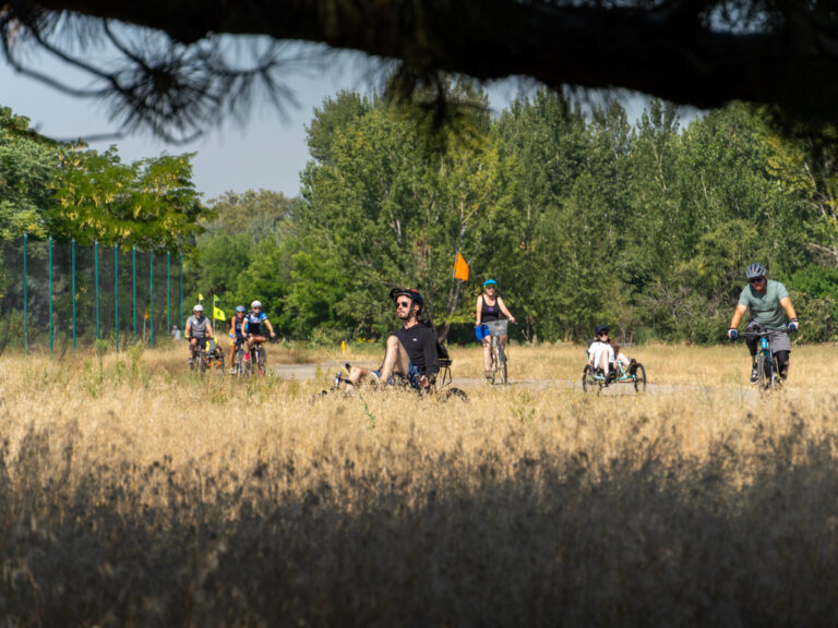 A group of cyclists, including adults and a child, ride along a dirt path surrounded by dry grass and green trees in a sunny outdoor setting.