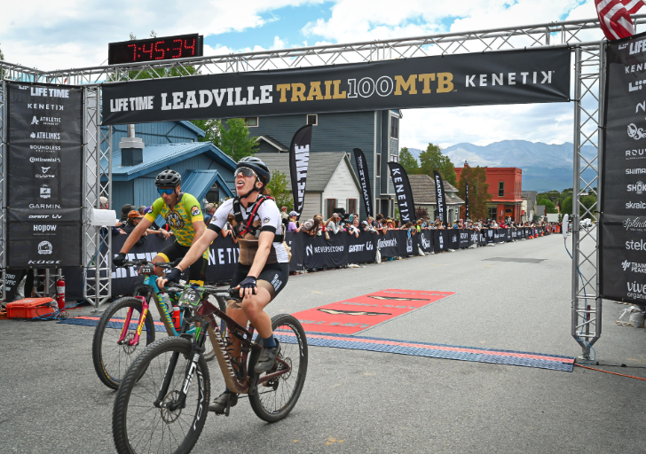 Adaptive cyclist crosses the finish line at the Leadville Trail 100 MTB race in Colorado, riding beneath the official race arch surrounded by spectators and mountain views.