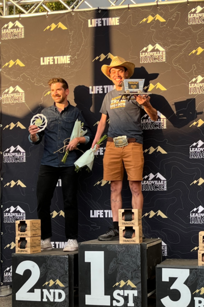 Athlete stands on the podium during the Leadville Trail 100 MTB awards ceremony, holding a trophy and race recognition plaque.