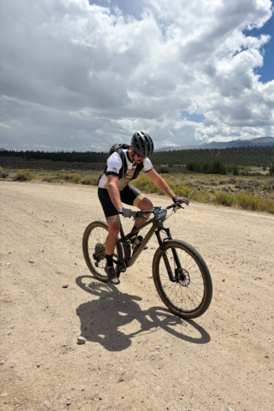Adaptive cyclist pedals along a gravel road during the Leadville Trail 100 MTB race, with Colorado mountains and dramatic clouds in the background.