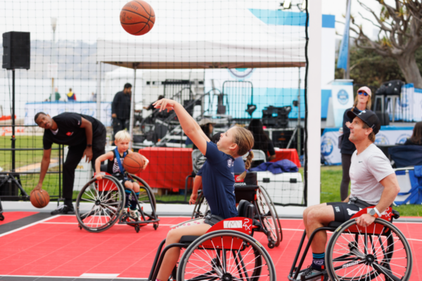 A young athlete in a sport wheelchair shoots a basketball during an adaptive basketball clinic while other athletes and coaches participate on the court in the background.