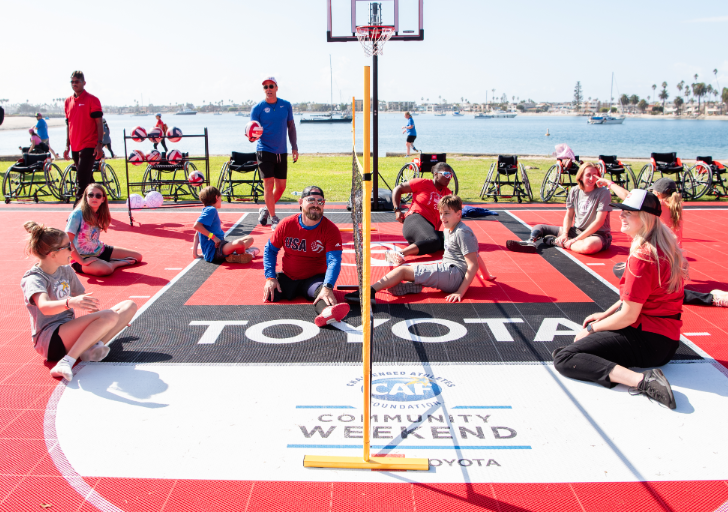 Athletes, coaches, and participants sit on an adaptive sport court during a CAF and Toyota Community Weekend activity beside the waterfront, with adaptive sports equipment and wheelchairs visible around the court.