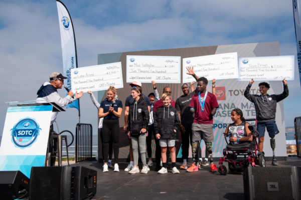CAF athletes stand onstage holding grant award checks during an SDTC presentation, while event speakers and supporters gather beneath a large video screen displaying Toyota branding.