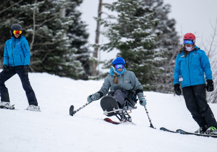 Three winter sports participants descend a snowy slope, including a person using an adaptive sit-ski in the center.