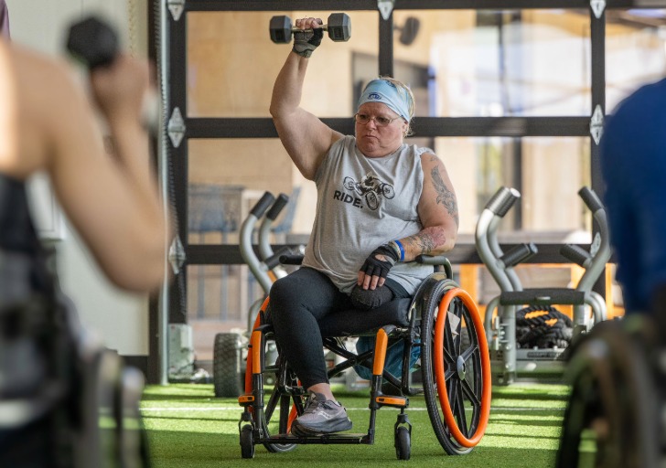 A person in a wheelchair lifts a dumbbell overhead in a brightly lit gym.