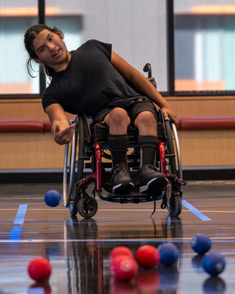 Person in a sports wheelchair reaching toward a blue ball on an indoor court, with red and blue balls scattered in the foreground.