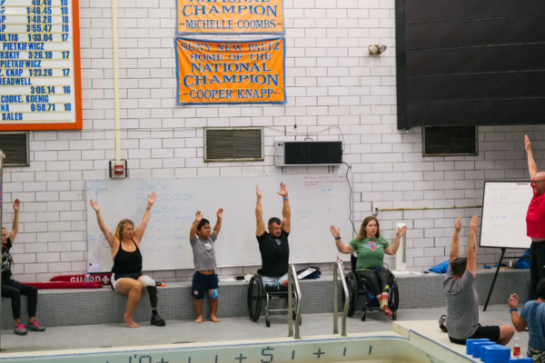 Adaptive athletes and clinic participants raise their arms during instruction beside the pool at the CAF Northeast Paratriathlon Clinic.