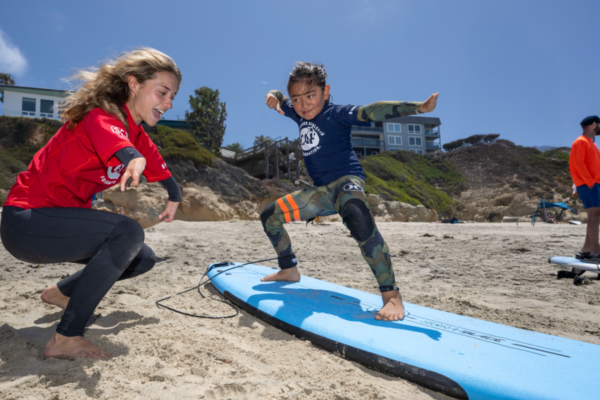 Adaptive surf instructor coaches a young participant practicing balance and surfing skills on a surfboard during a beach clinic.