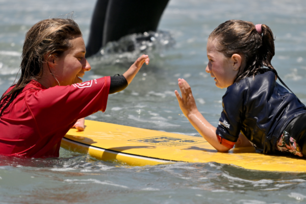 Adaptive surf instructor and youth participant share a high-five while practicing surfing skills in the ocean during a CAF adaptive surf clinic.