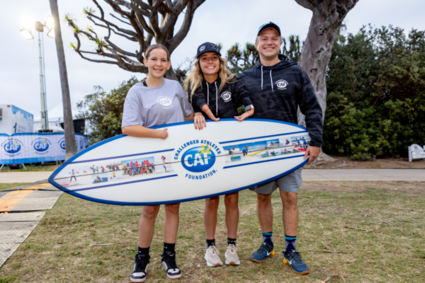 Youth participant stands with family members while holding a CAF-branded surfboard at an adaptive surfing event.