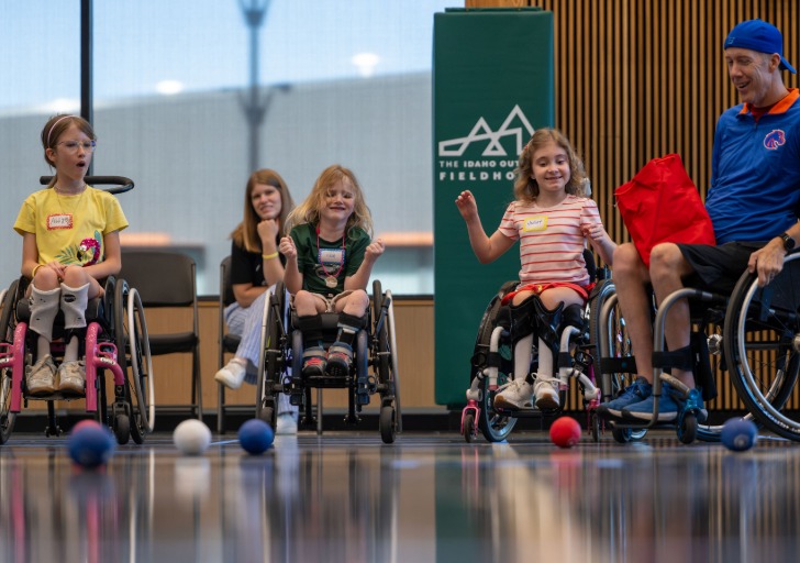 Children and an adult in wheelchairs participate in adaptive bowling indoors, with colorful bowling balls on the polished floor.