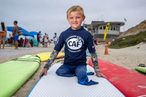 Young Challenged Athletes Foundation participant sits on a surfboard during an adaptive surfing clinic at the beach.