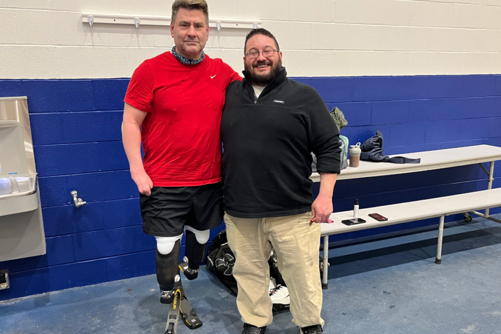 1. Jeff Belcher, wearing lower-limb prostheses, stands with a friend at an indoor sports facility.