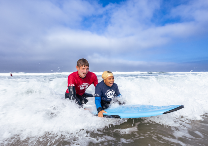 Young adaptive athlete rides a wave on a surfboard with assistance from a CAF volunteer during an adaptive surfing experience.