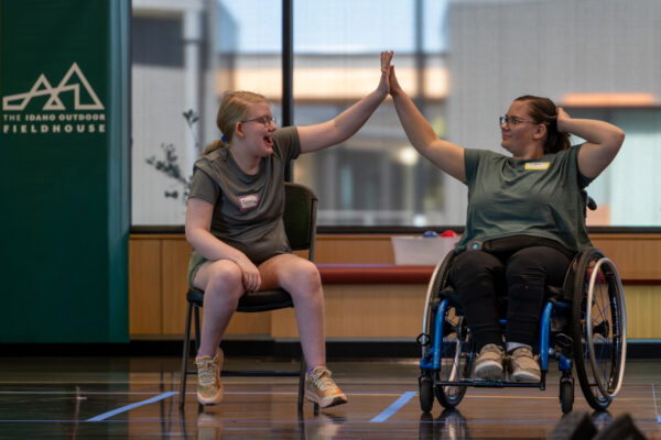 Two people high-five while seated in a chair and a wheelchair on an indoor court.