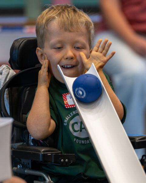 Smiling child in a wheelchair raises both hands beside their face as a blue ball rests on a white ramp in front of them.