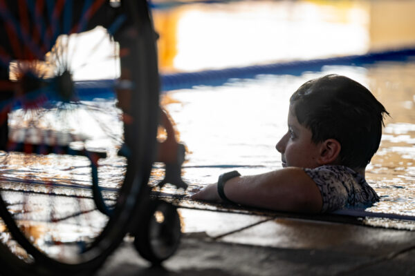 Swimmer with wet hair resting on the edge of a sunlit indoor pool, partially obscured by blurred equipment in the foreground.