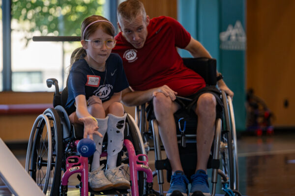 Young girl in a wheelchair reaches toward a blue ball while an adult man in another wheelchair sits behind her in an indoor gym.